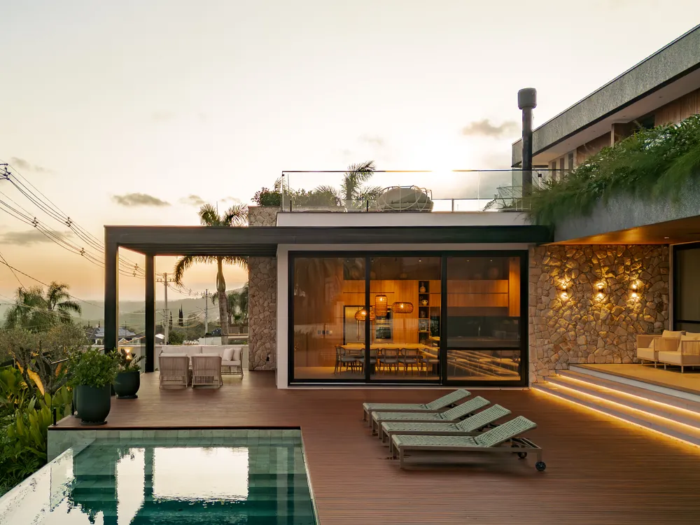 Residência ao anoitecer com porta de correr de piso a teto, cozinha acesa ao fundo, deck de madeira e piscina
